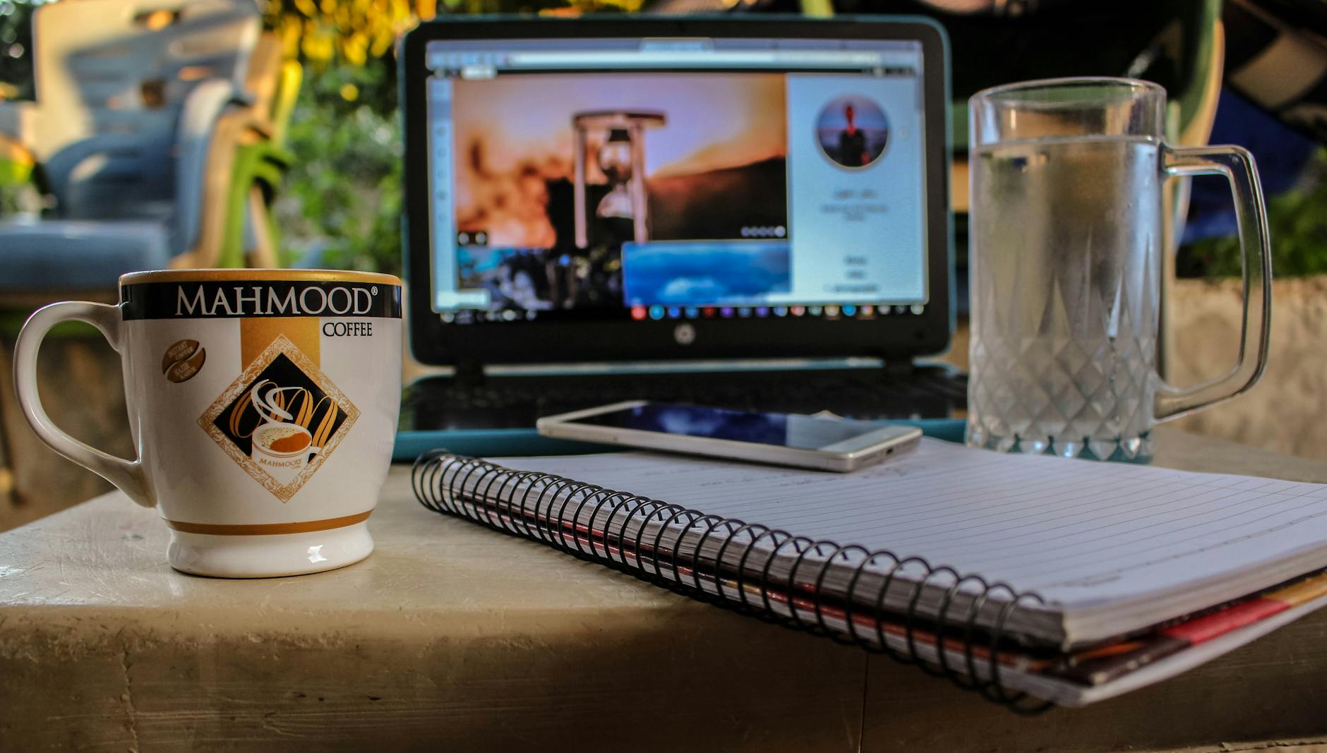 white smartphone on spiral notebook between ceramic mug and glass beer mug near black computer monitor showing hour glass