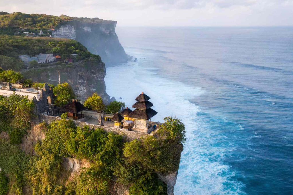 Aerial view of a cliffside temple in Bali, surrounded by lush greenery and overlooking the ocean waves.