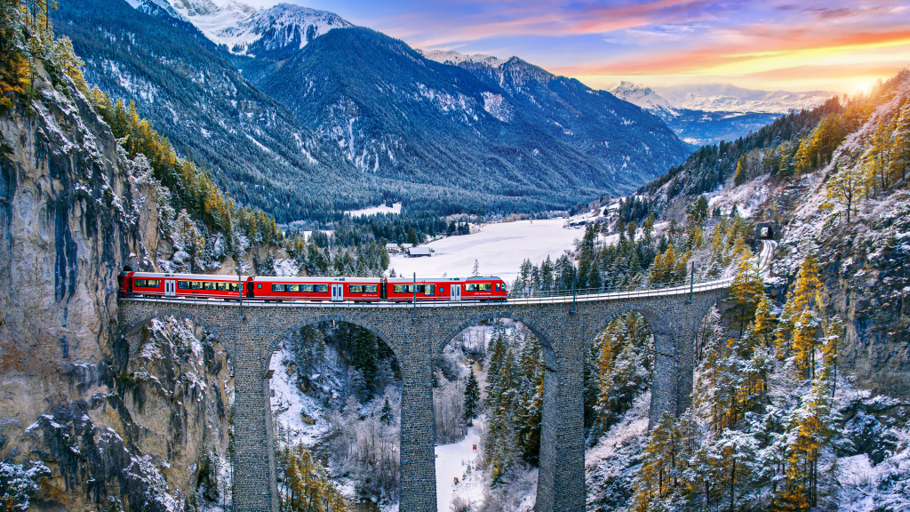 A scenic view of a red train crossing a stone bridge over a snow-covered valley, surrounded by lush green forests and majestic mountains during sunset.